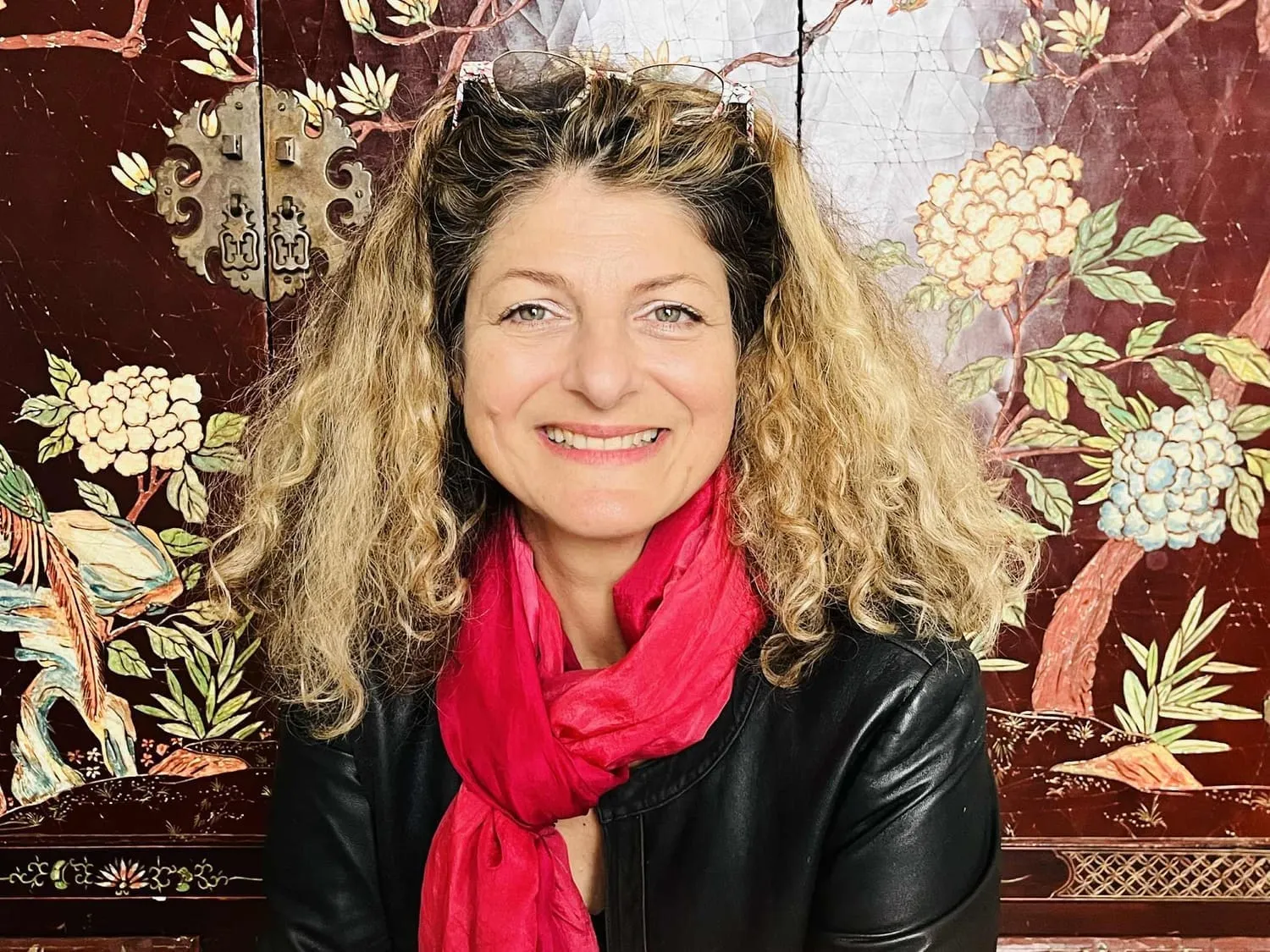 Portrait souriant d'une femme aux cheveux bouclés blonds, foulard rose vif et veste en cuir noir, devant un panneau laqué décoré de fleurs