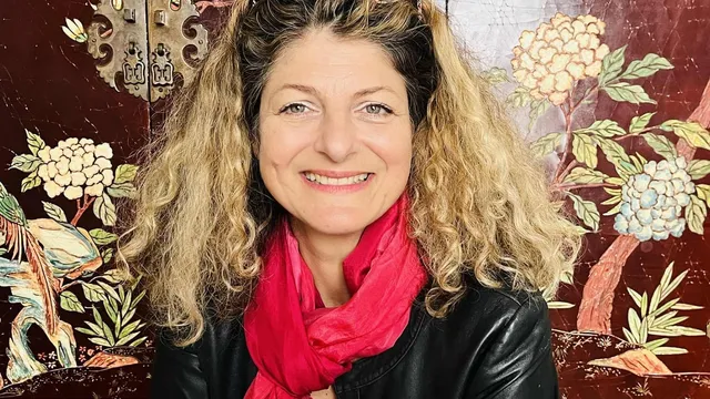 Portrait souriant d'une femme aux cheveux bouclés blonds, foulard rose vif et veste en cuir noir, devant un panneau laqué décoré de fleurs